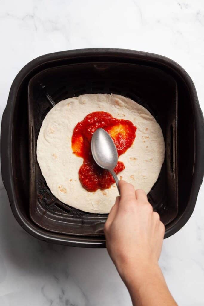 spreading marinara sauce on flour tortilla in an air fryer basket