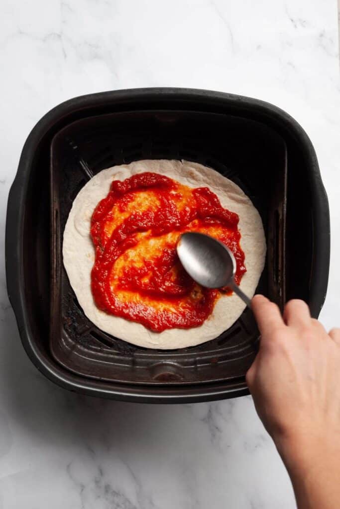 adding red sauce on a tortilla in an air fryer basket before adding cheese